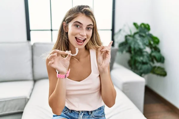 Young blonde woman holding invisible aligner orthodontic with a big smile on face, pointing with hand finger to the side looking at the camera.