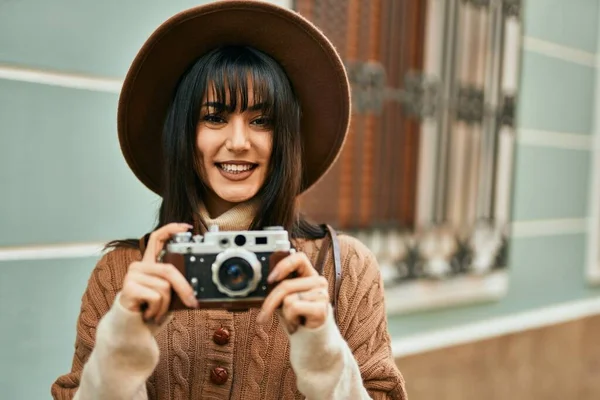 Brunette woman wearing winter hat smiling using vintage camera outdoors at the city
