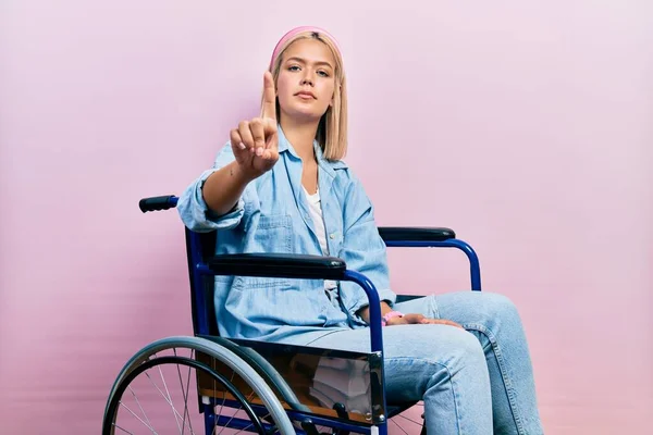 Красивая Блондинка Сидящая Инвалидном Кресле Показывая Пальцем Вверх Сердитым Выражением — стоковое фото