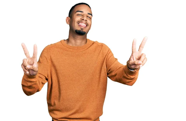 両方の手の指を示す舌で笑顔カジュアルな服を着ている若いアフリカ系アメリカ人の男性は勝利のサインを行う — ストック写真