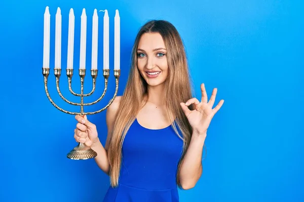 Młoda Blondynka Gospodarstwa Menorah Hanukkah Świeca Żydowska Robi Znak Palcami — Zdjęcie stockowe