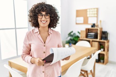 Ofiste iş elbisesi giyen genç bir ortadoğulu kadın yüzünde mutlu ve havalı bir gülümsemeyle. şanslı kişi. 