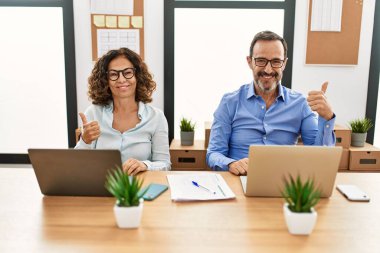 Orta yaşlı İspanyol bir kadın ve ofiste dizüstü bilgisayarlı bir adam el hareketleriyle mutlu mesut el hareketi yapıyorlar. Kameraya bakıp başarıyı gösteren ifade. 