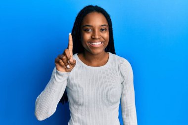 Saçları örgülü beyaz süveter giyen Afro-Amerikalı bir kadın kendini beğenmiş ve mutlu bir şekilde gülümserken bir numarayı işaret ediyor.. 