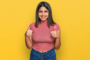 Günlük tişört giyen genç İspanyol kız. Başarı işareti ile el hareketi yapıyor. Gülümsüyor ve mutlu. Neşeli ifade ve kazanan jest. 