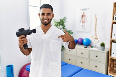 Genç Hintli fizyoterapist elinde masaj tabancasıyla Sağlık Merkezi 'ne bakıyor. Yüzünde bir gülümsemeyle, kendini gururlu ve mutlu olarak gösteriyor.. 