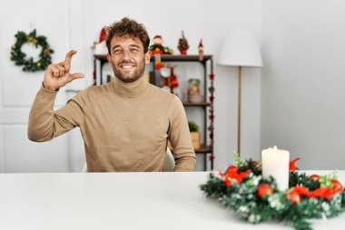 Genç, sakallı yakışıklı bir adam Noel süslemesi masasında oturuyor gülümsüyor ve el hareketleriyle küçük boy işareti yapıyor, parmakları görünüyor ve kameraya bakıyor. ölçüm kavramı. 