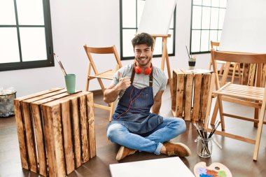 Sanat stüdyosunda oturan İspanyol genç adam elleriyle mutlu bir jest yapıyor. Kameraya bakıp başarıyı gösteren ifade. 