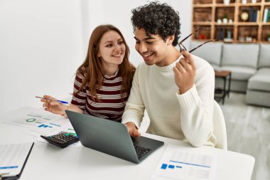 Genç çift evde masada dizüstü bilgisayar kullanarak mutlu bir şekilde gülümsüyor..