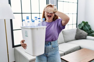 Genç kızıl saçlı kadın elinde plastik şişelerle çöp sepetini tutarken gülümsüyor ve elleri yüzünde şaşkınlıktan gözleri kapatıyordu. kör kavram. 