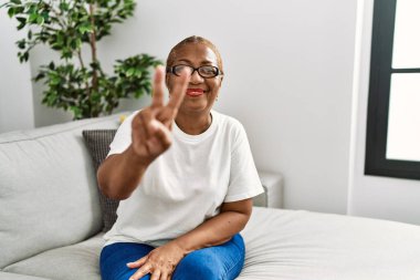 Olgun İspanyol kadın evdeki kanepede oturmuş kameraya bakıp zafer işareti yapıyor. İki numara.. 