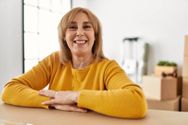 Orta yaşlı sarışın kadın mutlu bir şekilde gülümsüyor. Yeni evindeki karton kutuya yaslanıyor..