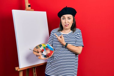 Orta yaşlı İspanyol kadın paletle resim çiziyor. Resim sehpası bir yana endişeli ve endişeli bir şekilde işaret parmağı, endişeli ve şaşırmış bir ifade sergiliyor. 