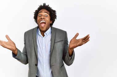 Genç Afro-Amerikan adam iş ceketi giyerek izole edilmiş beyaz arka planda zaferi mutlu bir gülümsemeyle ve ellerini kaldırarak kazanan ifadeyle kutluyor. 