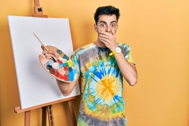 Genç, İspanyol asıllı bir adam boya sehpası üzerine palet çizerek ayakta dikiliyor. Ağzını yanlışlıkla ellerle kapatıyor. gizli kavram. 