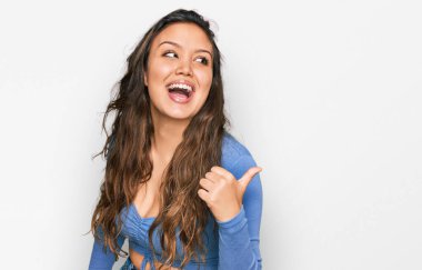 Gündelik kıyafetler giyen, gülümseyen, gülümseyen ve başparmağıyla yan tarafı gösteren İspanyol genç kız.. 