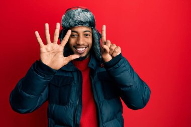 Sakallı, kışlık şapkalı ve paltolu genç Afrikalı Amerikalı adam altı numaralı parmaklarını gösterirken kendinden emin ve mutlu bir şekilde gülümsüyor.. 