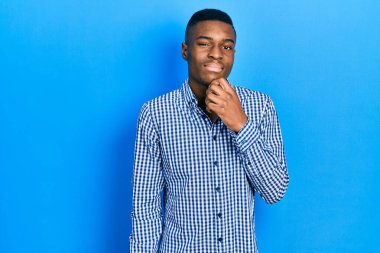 Gündelik giysiler giymiş genç Afro-Amerikan adam kameraya kendinden emin bakıyor kollarını kavuşturmuş ve çenesini kaldırmış bir şekilde gülümsüyor. Olumlu düşünmek. 