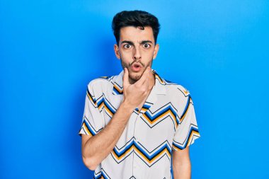 Gündelik kıyafetler giyen genç İspanyol adam, elleri çenesinde, inanamama, şaşkınlık ve şaşkınlık dolu bir ifade takınıyor. 