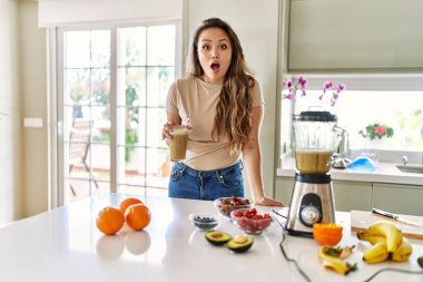 Güzel, genç, esmer bir kadın mutfakta bir bardak smoothie içiyor. Korkmuş ve ağzı açık bir şekilde şaşkınlık içinde. 