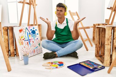 Sanat stüdyosundaki genç İspanyol adam, elleri ve kolları havada, kafası karışık bir ifade takınıyor. şüphe kavramı. 