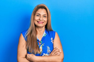 Orta yaşlı İspanyol kadın gündelik kıyafetler giyip kollarını kavuşturup kameraya bakarken gülümsüyor. pozitif kişi. 