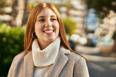 Genç kızıl kız gülümsüyor, mutlu bir şekilde şehirde duruyor..