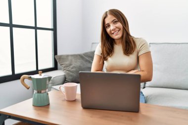 Genç esmer kadın evde dizüstü bilgisayar kullanıyor. Bir fincan kahve içiyor. Kollarını kavuşturup kameraya bakıyor. pozitif kişi. 