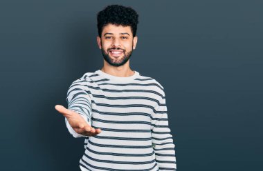 Sakallı, sıradan çizgili süveter giyen neşeli gülümseyen, yardım eli uzatan ve kabul gören genç bir Arap.. 