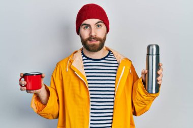 Sakallı, elinde kahve fincanı ve termosu olan beyaz bir adam, ağzı açık kalmış, şüpheci ve alaycı görünüyor. 