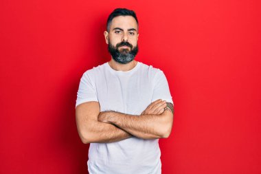 Gündelik beyaz tişört giyen genç İspanyol adam kameraya bakarken kollarını kavuşturup gülümsüyor. pozitif kişi. 