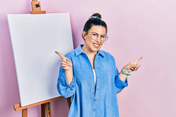 画家のイーゼルのそばに立つ若いヒスパニック系の女性は さまざまな方向に指で自信を持って指して笑顔立っている 広告のコピースペース — ストック写真