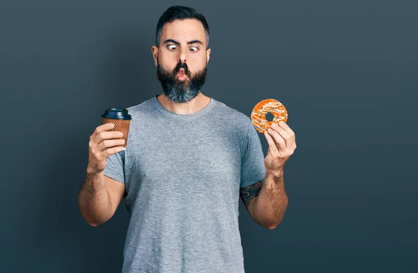 Hispanic man with beard eating doughnut and drinking coffee making fish face with mouth and squinting eyes, crazy and comical.