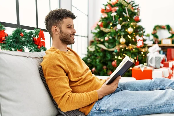 Young arab man reading book sitting on the sofa by christmas tree at home.
