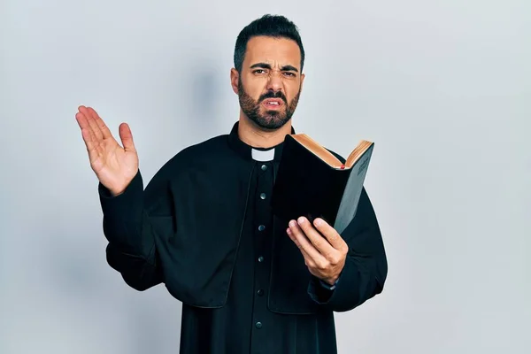 ひげを生やして聖書とクリスチャンの十字架の無知で混乱した表現を持つハンサムなヒスパニック系の司祭の男 疑わしい概念 — ストック写真
