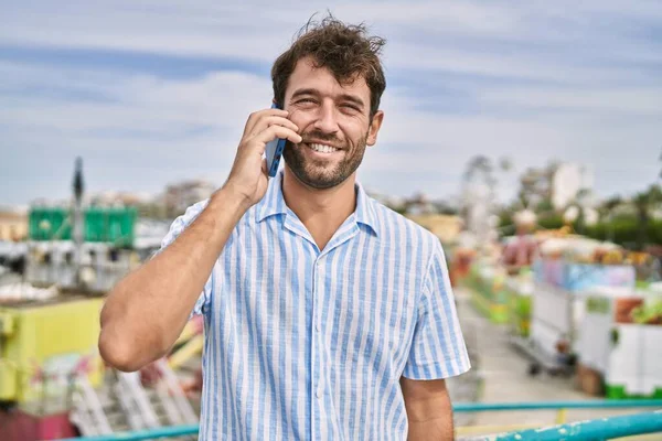 Genç İspanyol adam gülümsüyor. Eğlence parkında akıllı telefondan konuşuyor..