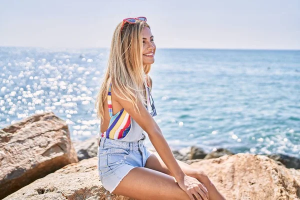 年轻的金发姑娘高兴地坐在海滩上的岩石上 — 图库照片