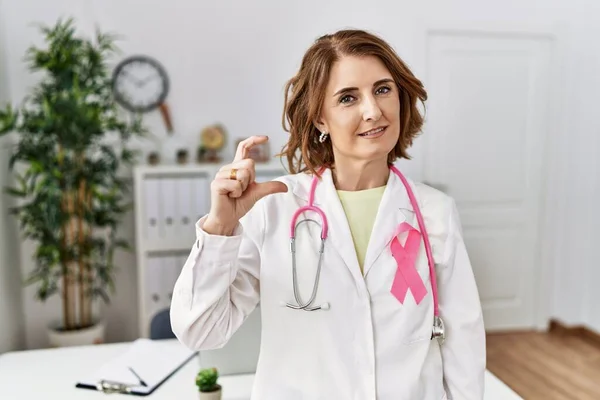 Middle age doctor woman wearing pink cancer ribbon on uniform smiling and confident gesturing with hand doing small size sign with fingers looking and the camera. measure concept.