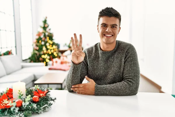 Joven Hombre Hispano Sentado Mesa Por Árbol Navidad Mostrando Señalando —  Fotos de Stock