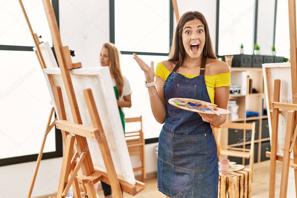 Young hispanic woman at art classroom celebrating victory with happy smile and winner expression with raised hands 