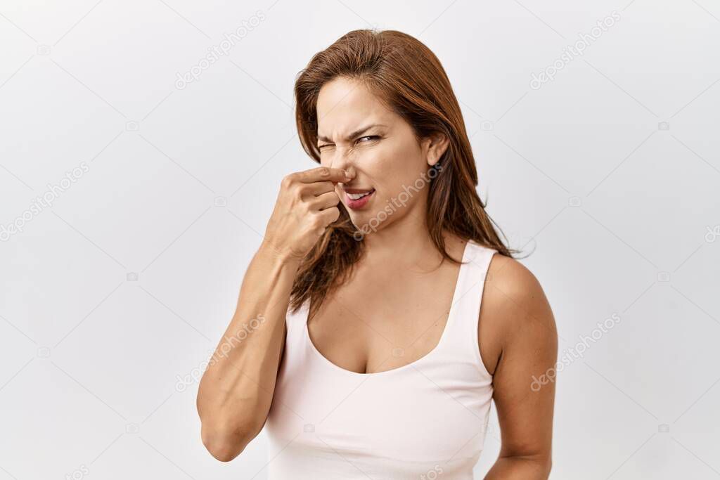 Middle age hispanic woman standing over isolated background smelling something stinky and disgusting, intolerable smell, holding breath with fingers on nose. bad smell 