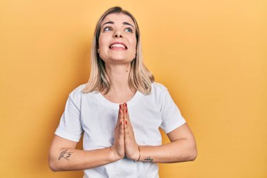 Güzel beyaz tişört giyen beyaz bir kadın elleri bağlı yalvarıyor ve dua ediyor. Yüzünde umut ifadesi var. Çok duygusal ve endişeli. Yalvarıyor. 