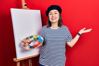 Orta yaşlı İspanyol kadın paletle resim çiziyor, resim sehpası ayakta gülümsüyor, eli kameraya bakıyor ve gülümsüyor.. 