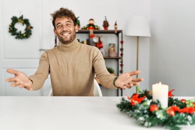 Masada sakallı yakışıklı genç adam Noel süslemeleri ile oturuyor. Neşeli gülümsüyor, kollarını açıyor. Dostça karşılama, olumlu ve kendinden emin selamlar. 