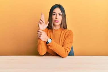 Latin kökenli genç bir kadın gündelik giysiler içinde masada oturmuş elinde sembolik bir silahla el hareketleriyle cinayet silahlarıyla oynuyor, kızgın bir yüz ifadesi takınıyor. 