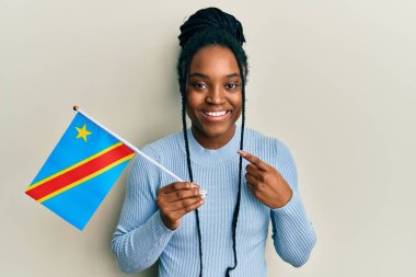 Saçları örgülü, Kongo bayrağının demokratik cumhuriyetini elinde tutan Afro-Amerikalı kadın mutlu bir şekilde elini ve parmağını göstererek gülümsüyor. 