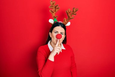 Young hispanic woman wearing deer christmas hat and red nose asking to be quiet with finger on lips. silence and secret concept.  clipart