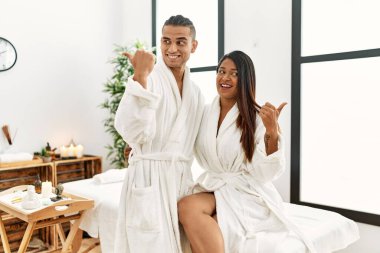 Young latin couple wearing towel standing at beauty center smiling with happy face looking and pointing to the side with thumb up. 