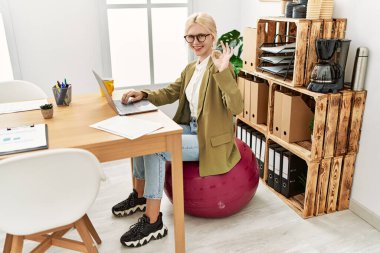 Ofiste çalışan beyaz, güzel bir iş kadını pilates topunda oturuyor. Parmaklarıyla imza atıyor, gülümsüyor. Mükemmel bir sembol. 