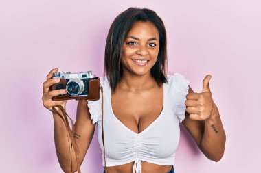 Afro-Amerikalı genç bir kız elinde klasik kamerayla mutlu ve pozitif gülümsüyor, başparmağıyla mükemmel ve onay işareti yapıyor. 